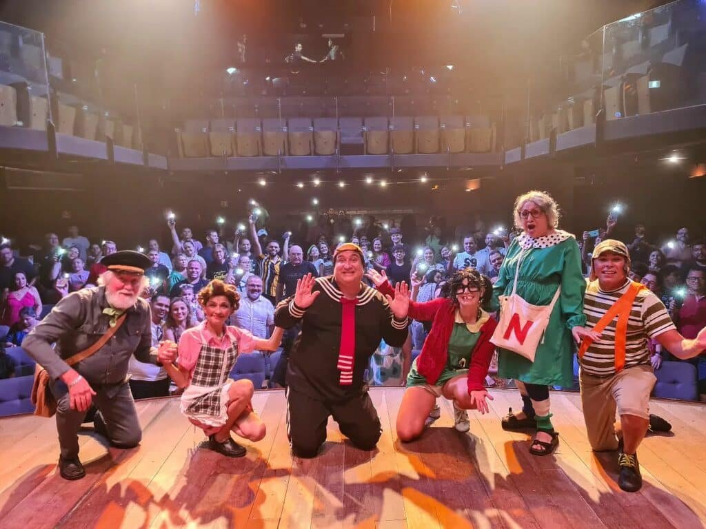 Foto de atores posando para a câmera com a plateia ao fundo durante um espetáculo no Teatro Brasil Tropical.