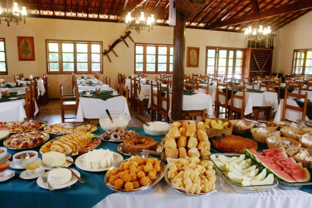 A imagem mostra um farto buffet de café da manhã em um ambiente rústico e acolhedor na Estalagem Fazenda Lazer. A mesa está repleta de pães, bolos, frutas frescas como melancia e melão, queijos, doces, roscas e outros quitutes tradicionais. Ao fundo, o salão é decorado com mesas cobertas por toalhas brancas e verdes, rodeadas por cadeiras de madeira. O espaço é bem iluminado, com janelas grandes que deixam a luz natural entrar, e o teto tem um estilo colonial com vigas de madeira e lustres.