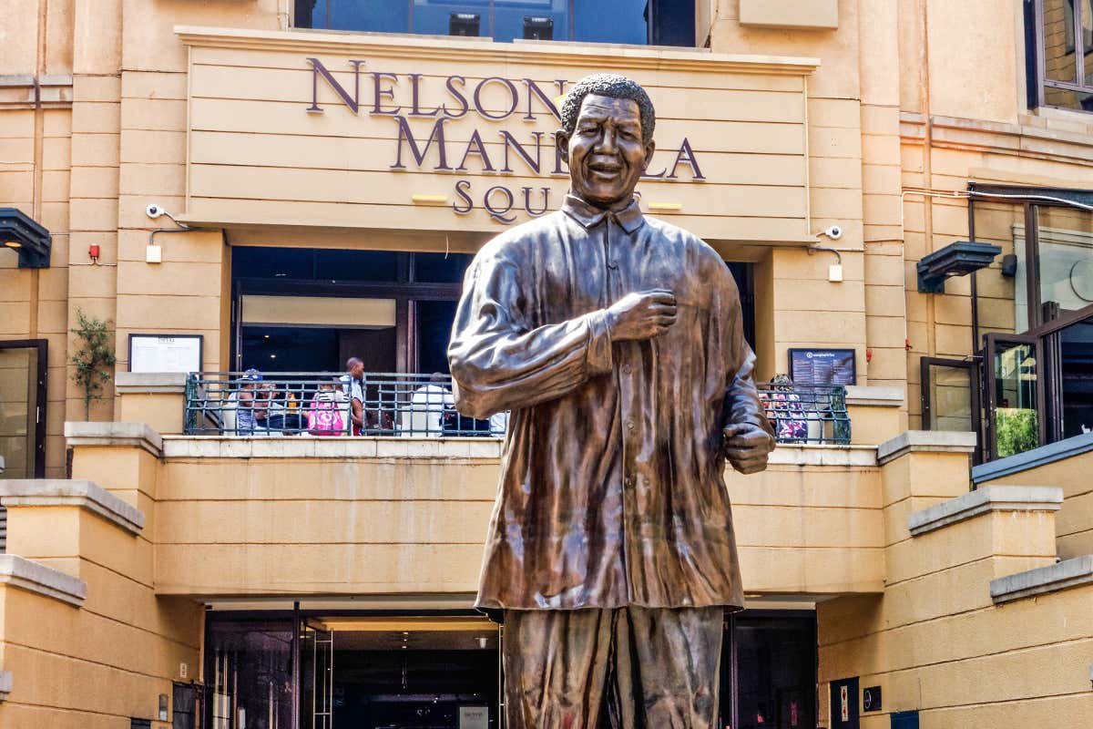 Uma alta estátua de bronze de um homem sorridente fica em frente a um edifício chamado "Praça Nelson Mandela", com varandas e janelas em Joanesburgo.
