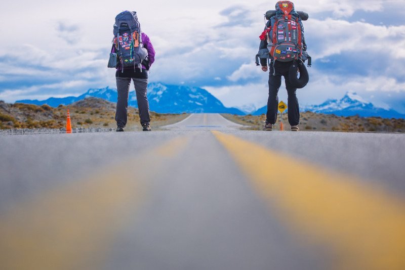 Dois mochileiros com mochilas grandes estão em uma estrada deserta, com montanhas e um céu nublado ao fundo. Imagem para ilustrar post sobre a Patagônia Argentina.