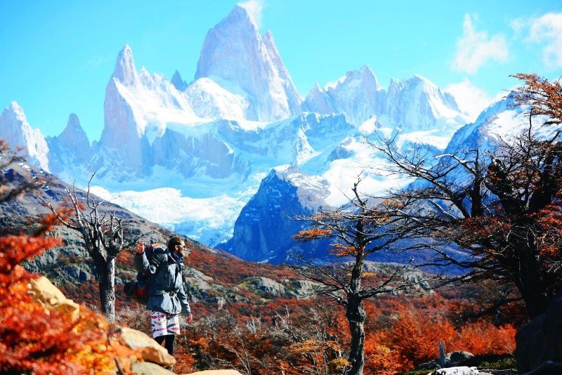 Uma pessoa com uma mochila está em um terreno rochoso, cercada por árvores de outono, com o Monte Fitz Roy ao fundo. Imagem para ilustrar post sobre a Patagônia Argentina.