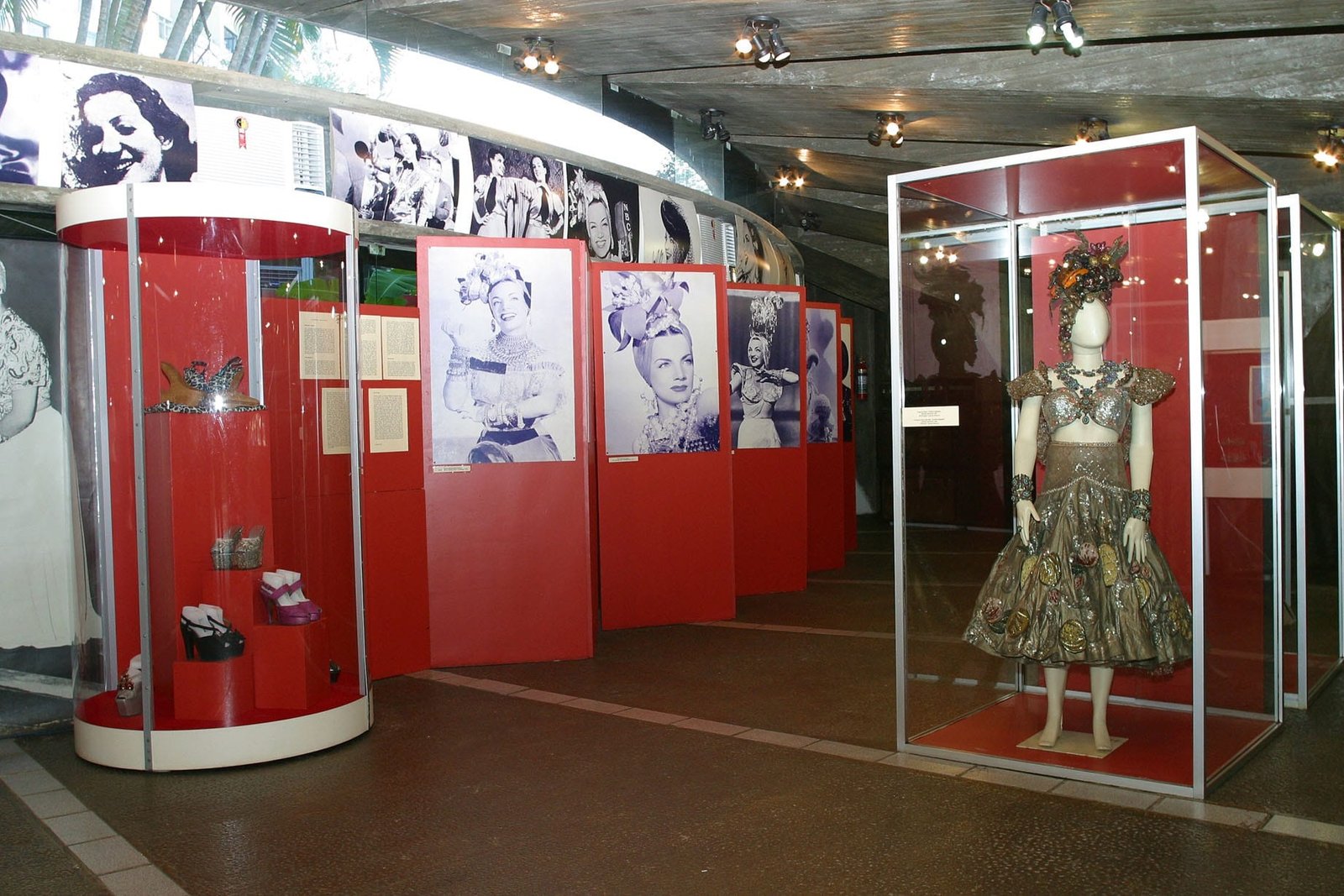 Exposição do Museu Carmen Miranda com painéis vermelhos com fotos em preto e branco, uma caixa cilíndrica com sapatos e um vestido em um manequim.