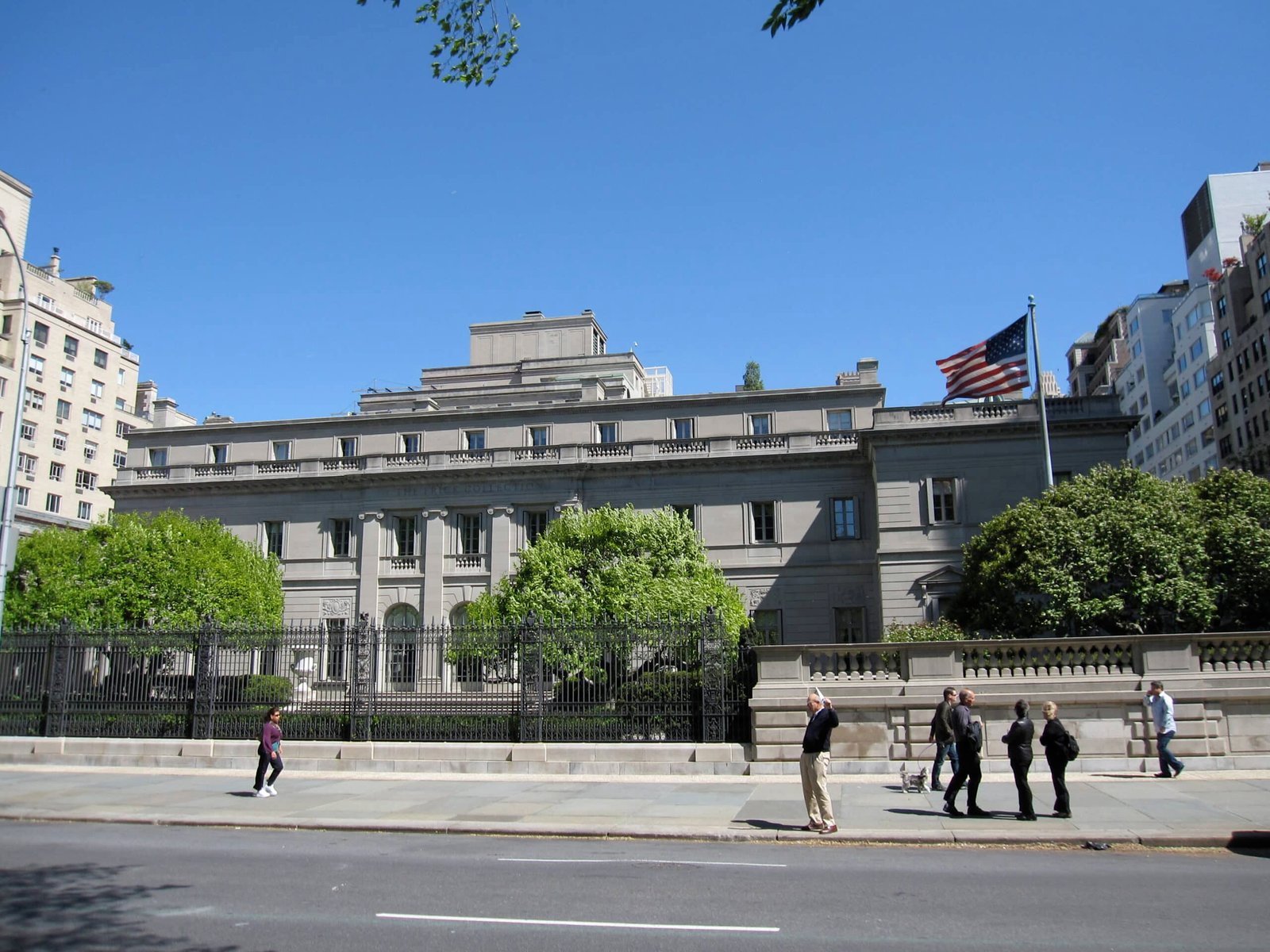 Fachada do Frick Collection com árvores e uma bandeira americana na frente. Várias pessoas estão caminhando na calçada.