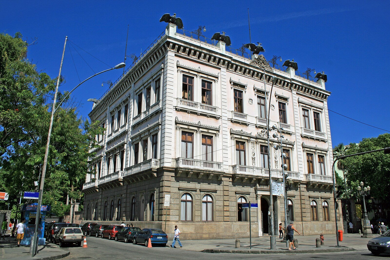 Palácio do Catete de três andares com decorações ornamentadas e estátuas no telhado, cercado por árvores e alguns pedestres.