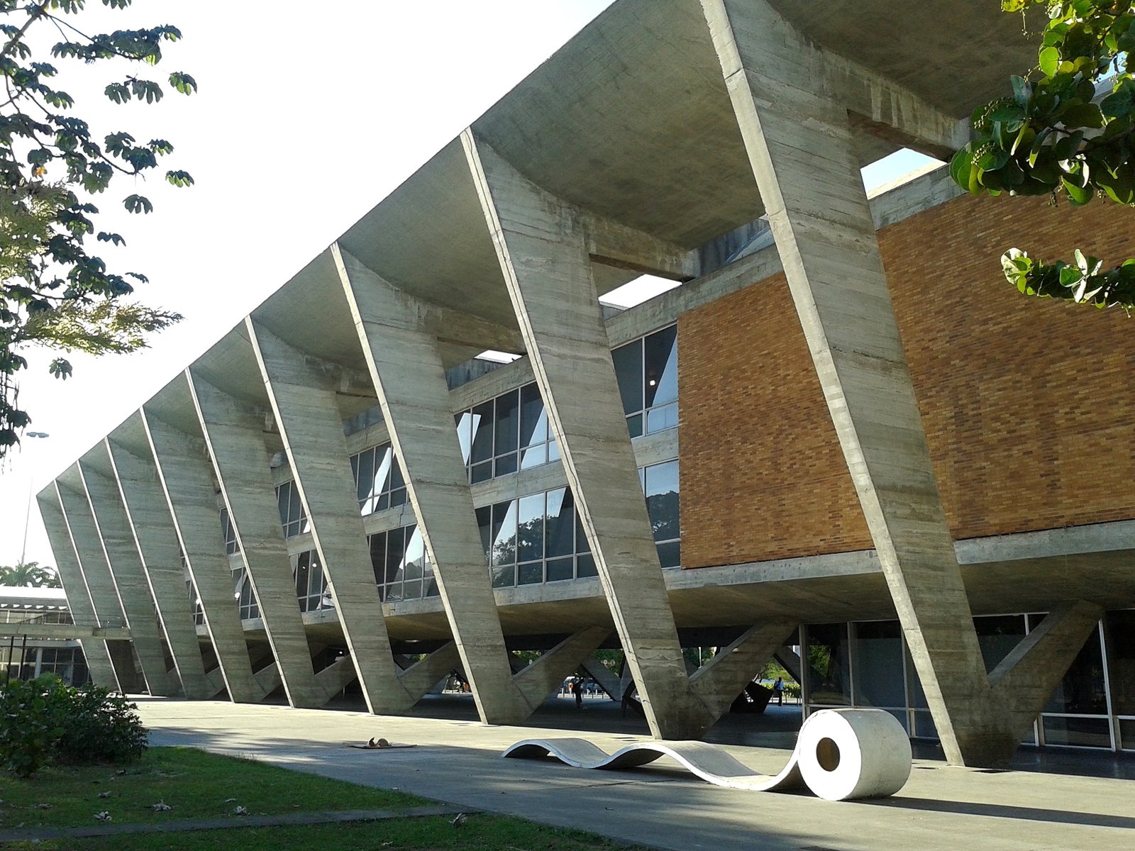 Museu de Arte Moderna com grandes colunas triangulares de concreto, paredes de tijolos e janelas de vidro. Uma escultura em forma de onda está em primeiro plano.