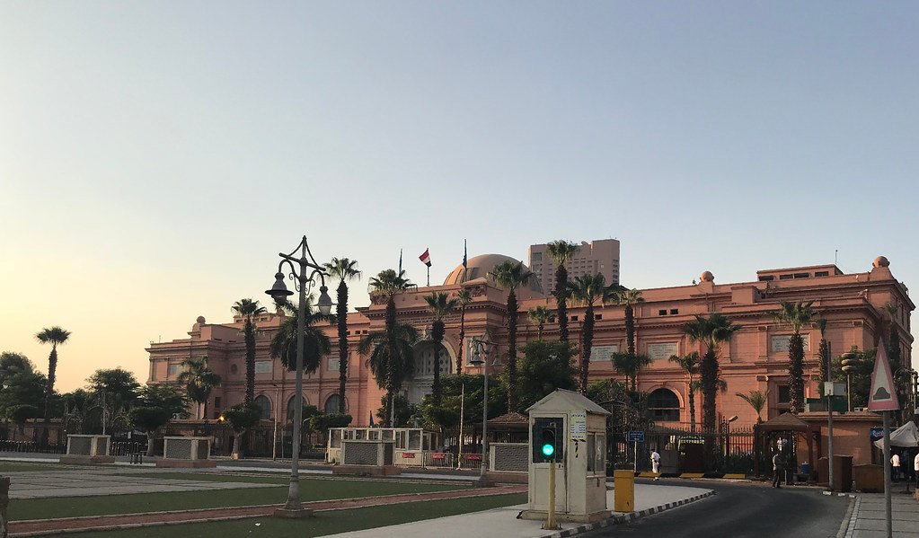 O Museu Egípicio em um grande edifício marrom-rosado com uma cúpula e bandeiras, cercado por palmeiras e postes de luz, ergue-se sob um céu claro.