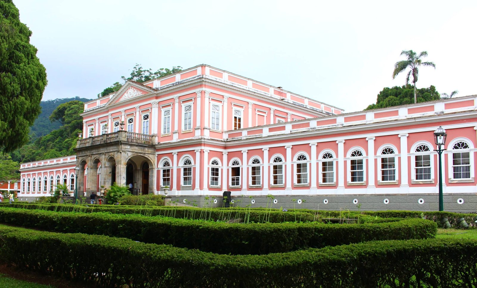 Museu Imperial rosa e branco se ergue com janelas em arco, cercado por sebes bem cuidadas e vegetação.