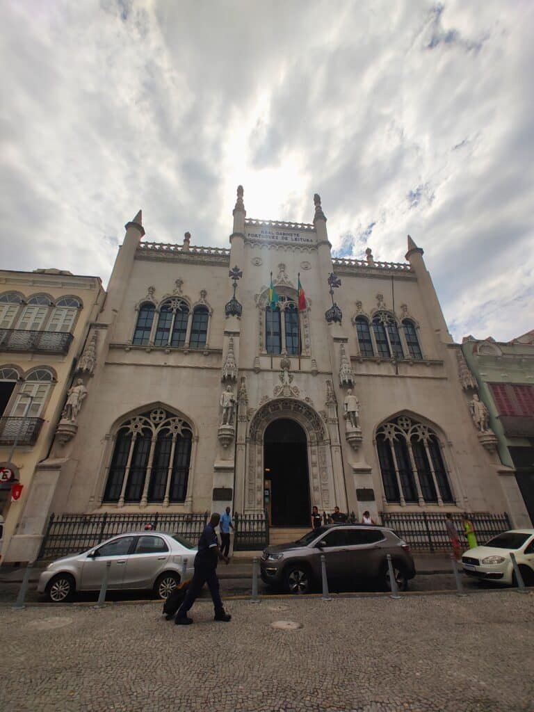 Edifício em estilo gótico com fachada ornamentada, bandeiras acima da entrada, carros estacionados e em movimento na frente e céu nublado no alto.