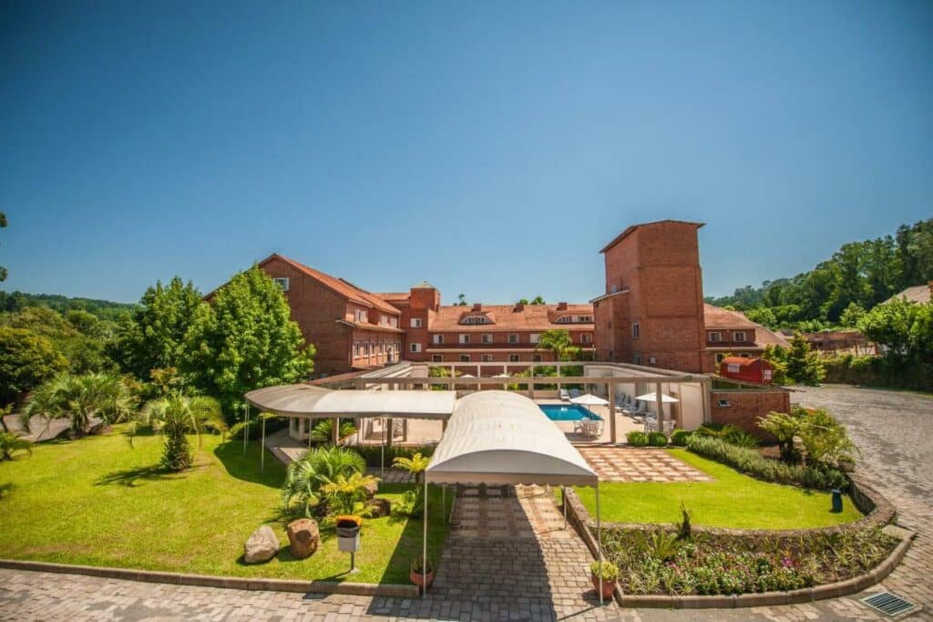 Vista da área externa do Farina Park Hotel, com gramado bem cuidado, árvores em crescimento, cobertura no caminho e piscina ao ar livre na frente do prédio do hotel