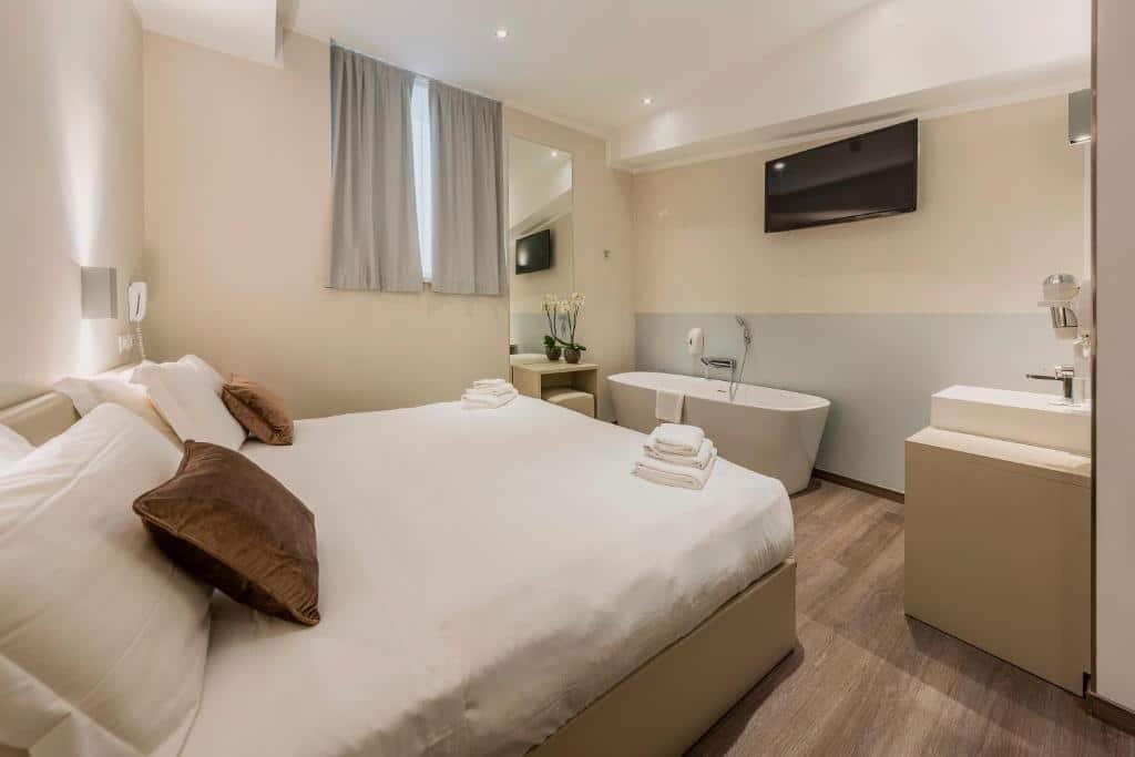 Quarto do Fasthotel Linate com uma cama de casal, uma janela com cortinas, um espelho de corpo inteiro, uma banheira oval perto de um móvel com uma pia, e há uma televisão na parede de frente para a cama, para representar hotéis perto do aeroporto de Milão