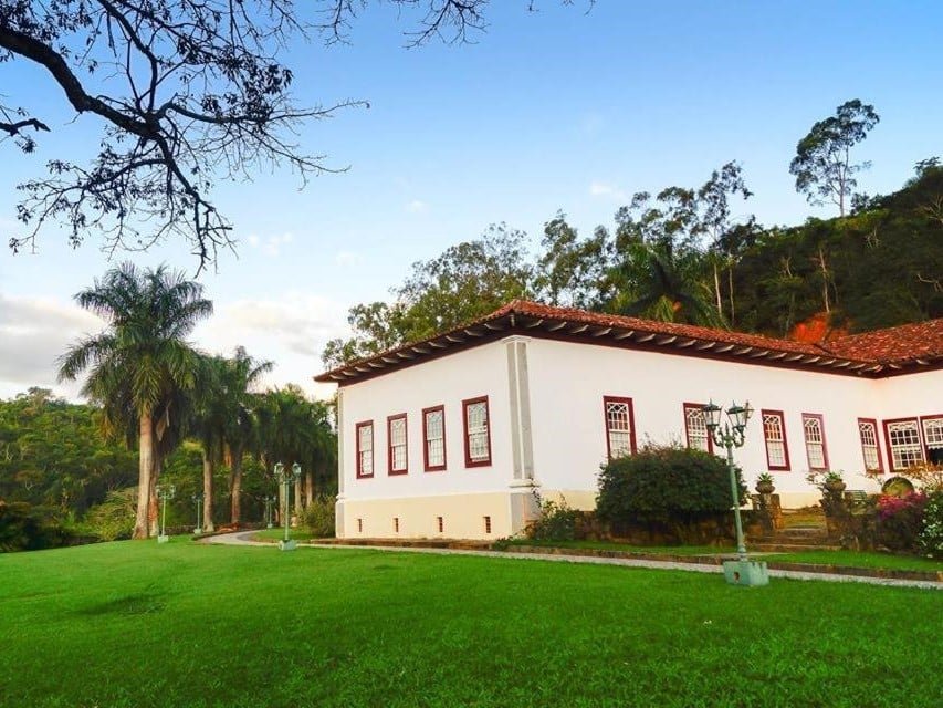 Edifício branco em estilo colonial com janelas com molduras vermelhas, cercado por vegetação exuberante e palmeiras sob um céu azul na Fazenda Cachoeira Grande, um dos hotéis fazenda no Rio de Janeiro.