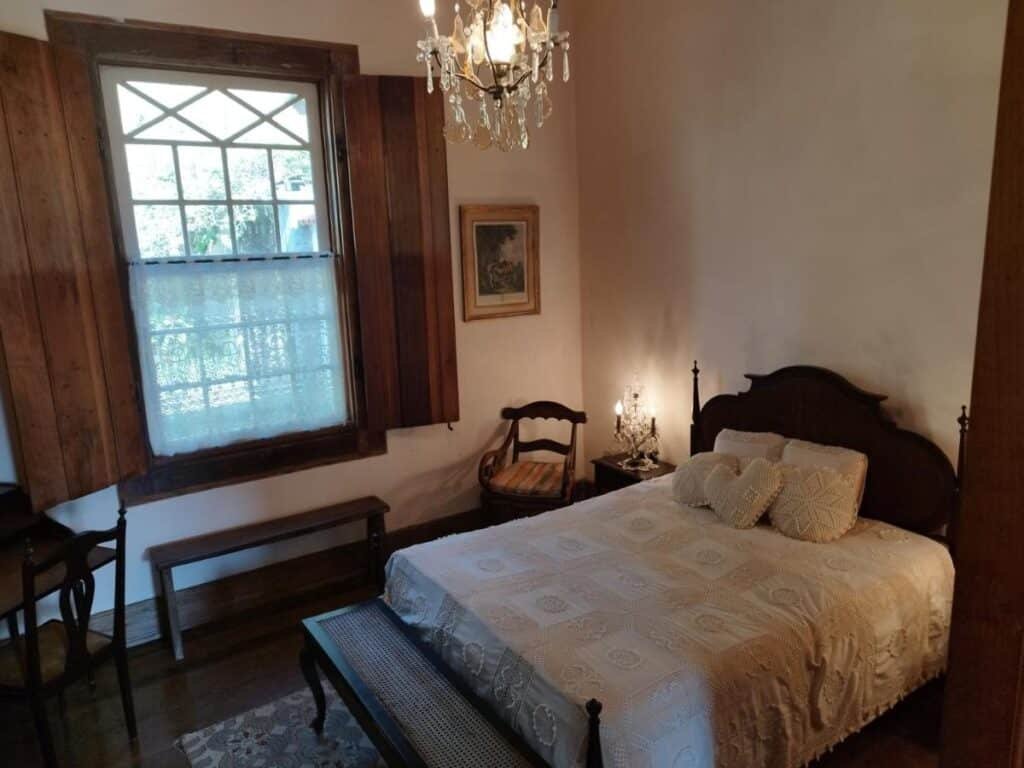 Um quarto da Fazenda Cachoeira Grande com uma cama de madeira vintage, colcha de renda, lustre, janela, venezianas de madeira, um pequeno banco e uma mesa lateral com um abajur.