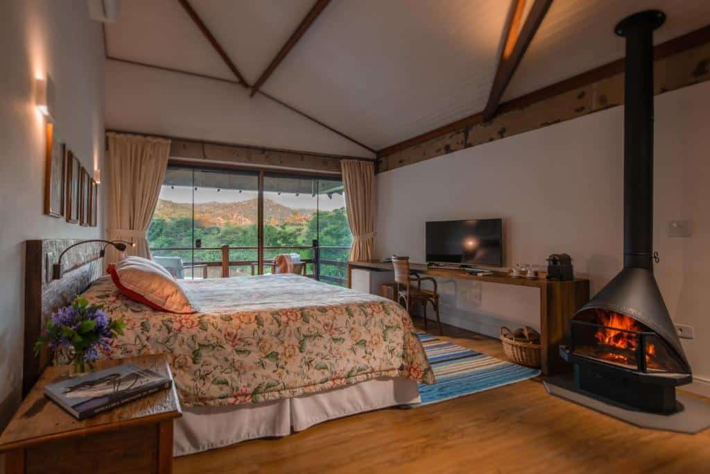 Um quarto aconchegante da Fazenda Capoava com uma cama king-size, roupa de cama floral, TV na parede, lareira e uma grande janela com vista para a vegetação.