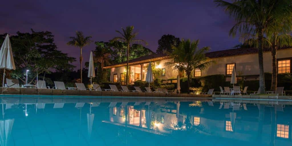 Vista noturna da fachada da Fazenda Capoava um dos hotéis fazenda em São Paulo iluminada com telhado de telhas e palmeiras, refletida em uma grande piscina cercada por espreguiçadeiras.