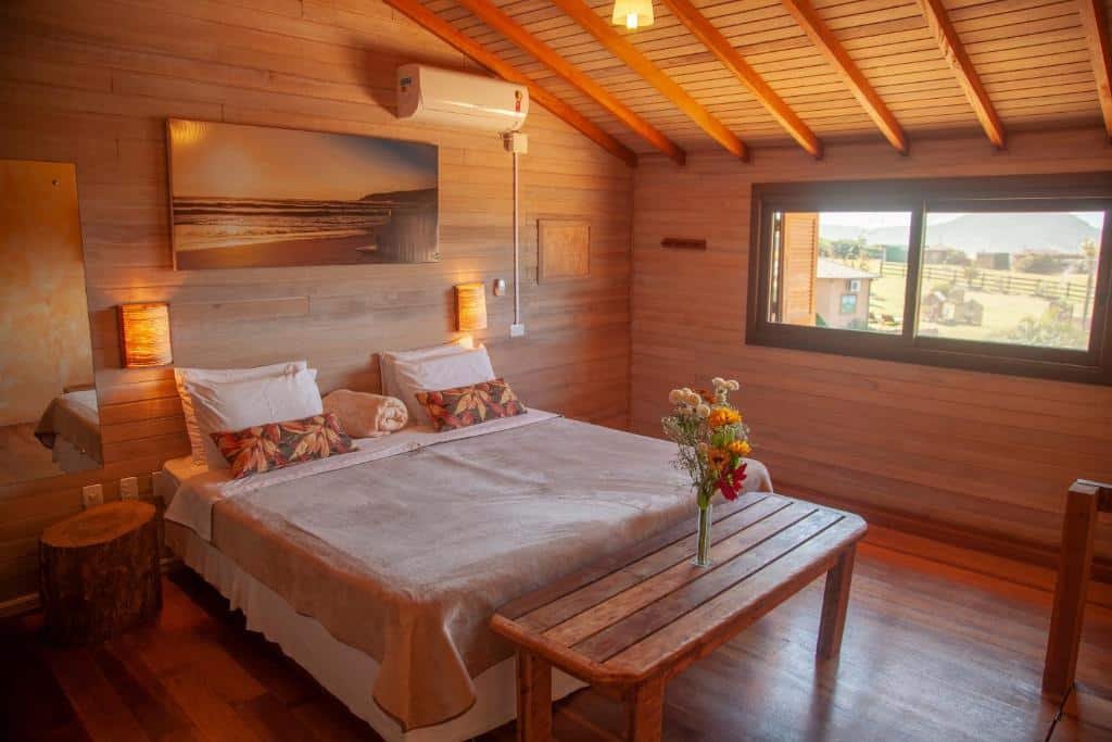 Um aconchegante quarto de madeira na Fazenda do Rosa com uma cama de casal, almofadas coloridas, um banco de madeira, uma janela com vista e um vaso de flores na mesa.