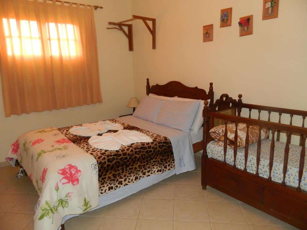 A imagem mostra um quarto aconchegante do Fazenda Hotel Alvorada com uma cama de casal e um berço de madeira ao lado. A cama de casal está coberta com uma colcha estampada de flores e uma manta com estampa de oncinha. Sobre a cama, há toalhas dobradas em formato decorativo. O berço de madeira combina com o estilo clássico da cama. A janela tem cortinas em tons de bege, deixando entrar luz suave. O quarto também possui decoração simples com quadros pequenos na parede, criando um ambiente acolhedor e familiar.