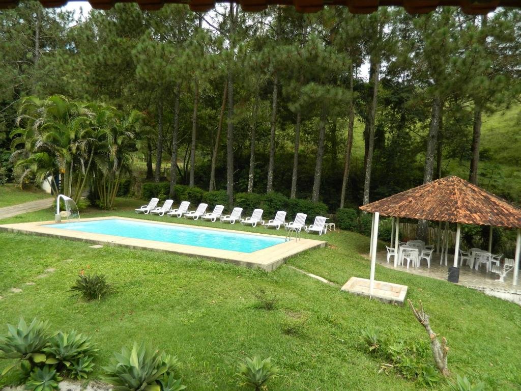 A imagem mostra uma área externa verdejante com uma piscina retangular cercada por cadeiras de sol brancas, organizadas em fila. Ao lado da piscina, há um pequeno quiosque com telhado de cerâmica, onde estão dispostas várias mesas e cadeiras brancas. A área ao redor é composta por um gramado verde, palmeiras e árvores altas que formam uma borda natural de pinheiros ao fundo, criando um ambiente tranquilo e acolhedor, perfeito para relaxar ao ar livre. Esse é o Fazenda Hotel Alvorada, um dos hotéis-fazenda em Minas Gerais.