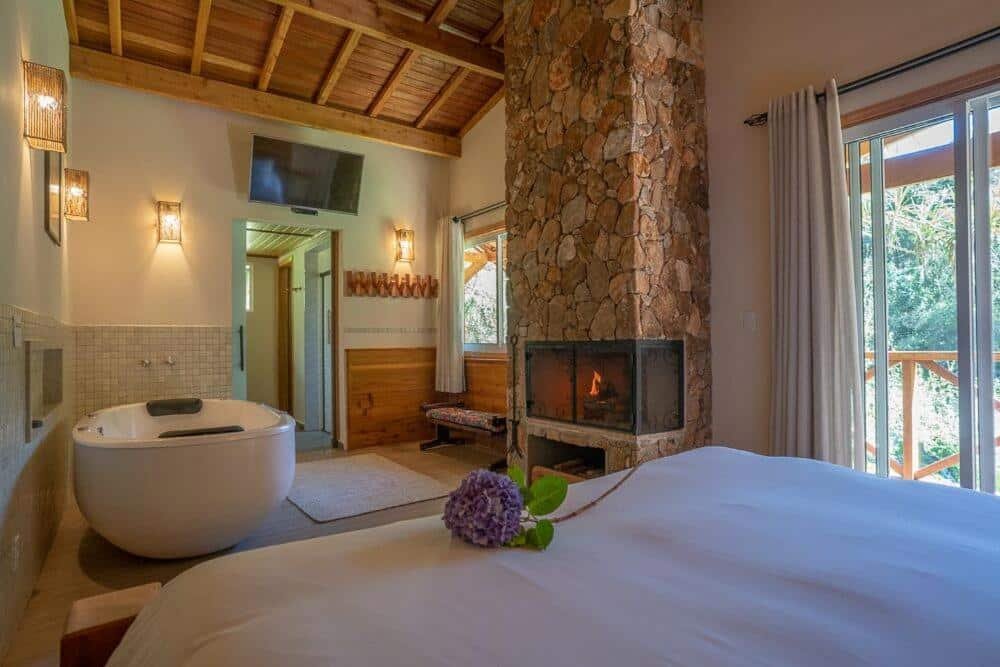 A imagem mostra um quarto aconchegante com uma decoração rústica e elegante do Fazenda Hotel Itapuá. No centro, há uma lareira de pedra acesa, ao lado de uma janela com cortinas claras que deixam entrar a luz natural e oferecem vista para a natureza externa. À esquerda, há uma banheira de hidromassagem moderna, próxima a uma parede decorada com ladrilhos suaves. Sobre a cama de casal, que ocupa o primeiro plano da imagem, há uma flor de hortênsia roxa colocada delicadamente. O teto é alto, com vigas de madeira expostas, criando um ambiente confortável e sofisticado.