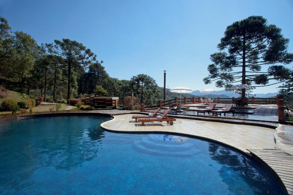 A imagem mostra uma bela piscina ao ar livre, cercada por espreguiçadeiras de madeira, com vista para uma paisagem montanhosa e coberta por árvores altas, incluindo araucárias. Ao lado da piscina, há um deck de madeira com guarda-sóis brancos, oferecendo sombra para algumas mesas e cadeiras. O céu está claro e azul, sem nuvens, e o ambiente é calmo e relaxante, ideal para aproveitar a natureza e a tranquilidade do local. A área ao redor da piscina é pavimentada, proporcionando um espaço confortável para os visitantes. Esse é o Fazenda Hotel Itapuá, um dos hotéis-fazenda em Minas Gerais.