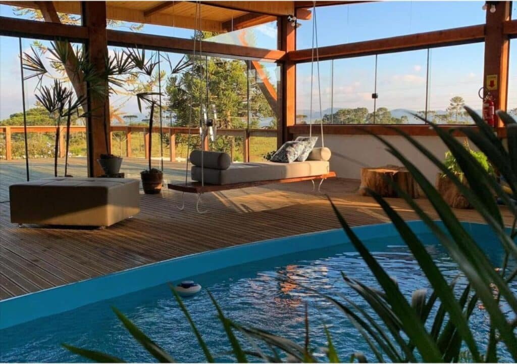 Deck de madeira interno com balanço, sofá e uma pequena piscina na Fazenda Santa Rita Turismo Rural. Grandes janelas revelam árvores e uma paisagem externa ensolarada.