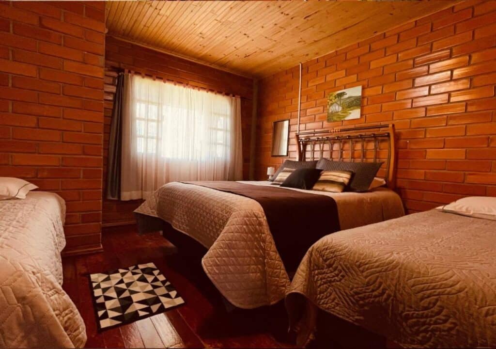 Um quarto aconchegante com uma cama de casal e duas camas de solteiro, todas com colchas, paredes de tijolos, uma janela com cortinas e teto e piso de madeira. Essa é a Fazenda Santa Rita Turismo Rural, um dos hotéis fazenda em Santa Catarina.