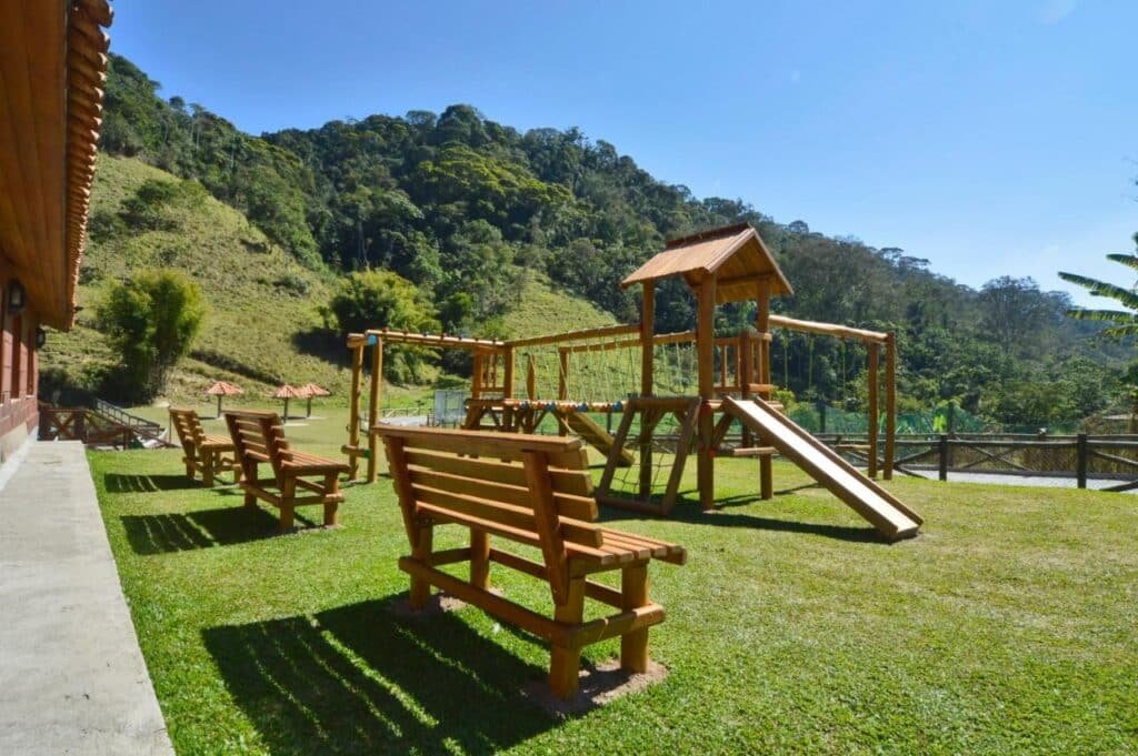Bancos de madeira e um playground em uma área gramada com colinas e árvores ao fundo, sob um céu azul claro na Fazenda Suíça Le Canton, um dos hotéis fazenda no Rio de Janeiro.