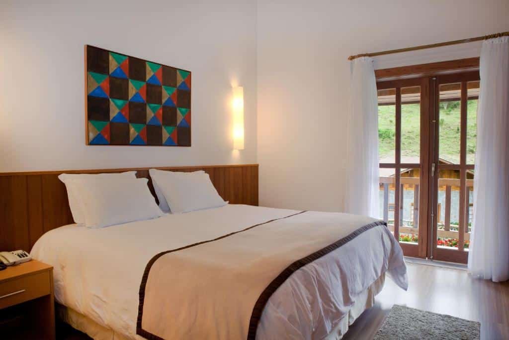 Um quarto aconchegante na Fazenda Suíça Le Canton com uma cama de casal bem arrumada, arte geométrica na parede, mesa de cabeceira e uma janela com vista para a vegetação.