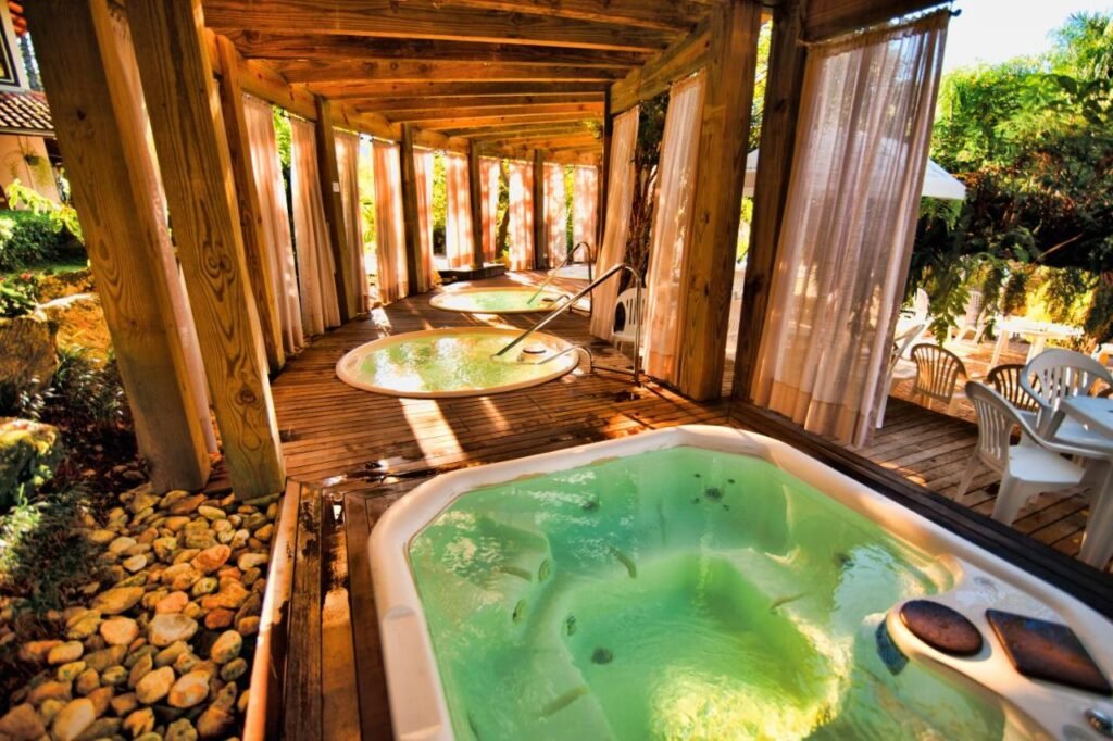 Área de spa ao ar livre do Fazzenda Park Resort com três banheiras de hidromassagem circulares em uma pérgola de madeira com cortinas. Duas cadeiras e uma mesa são visíveis ao fundo.