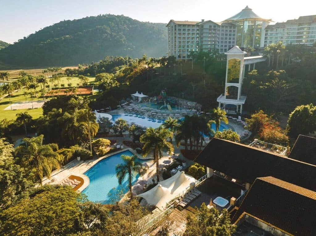 Vista aérea do Fazzenda Park Resort, um dos hotéis fazenda em Santa Catarina, com piscinas, palmeiras e prédios de vários andares, tendo como pano de fundo colinas e vegetação exuberante.