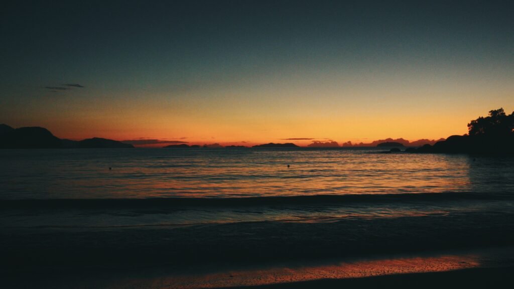 Uma vista serena do oceano ao pôr do sol com um brilho alaranjado no horizonte e ilhas silhuetas ao longe. Essa é a Praia do Félix em Ubatuba.