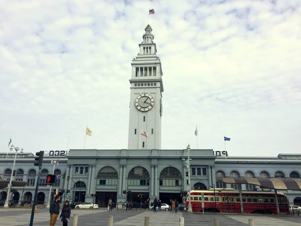 Vista de frente do Ferry Building. A torre do relógio fica bem no meio e em frente a construção passam pessoas, carros e um ônibus. A foto ilustra o post de o que fazer em San Francisco.