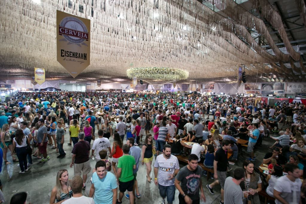 Vista de público do Festival Brasileiro da Cerveja, em galpão do Parque Vila Germânica. No teto existem bandeiras da cerveja EisenBahn e fitinhas decotativas. Foto do site oficial Parque Vila Germânica.