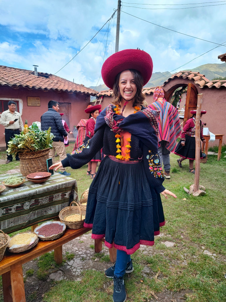 nátalie guimarães posando para a foto no centro da imagem. Ela usa as roupas típicas do vilarejo em Chinchero, no Vale Sagrado dos Incas: uma saia de lã preta com bordas vermelhas, um chapéu vermelho e um casaco azul com desenhos costurados.