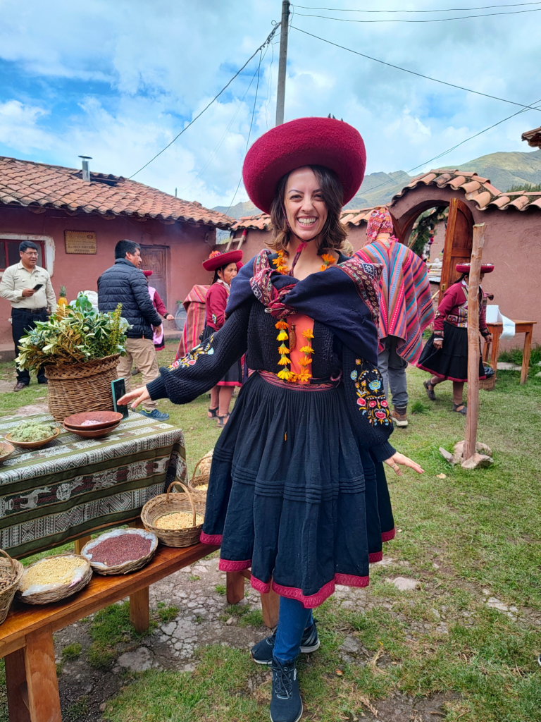 nátalie guimarães posando para a foto no centro da imagem. Ela usa as roupas típicas do vilarejo: uma saia de lã preta com bordas vermelhas, um chapéu vermelho e um casaco azul com desenhos costurados.