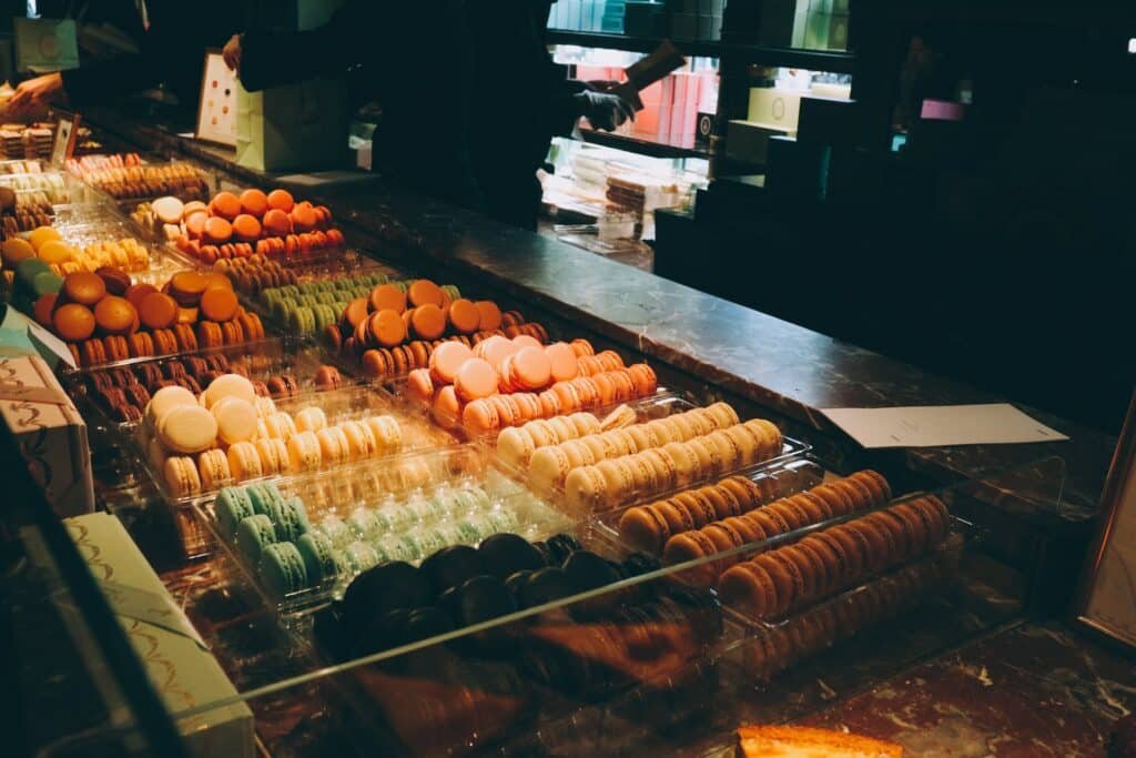 A imagem mostra uma vitrine da Ladurée repleta de macarons coloridos organizados em fileiras. Os macarons variam em cores, incluindo laranja, amarelo, verde, marrom e creme. Cada fileira está disposta em bandejas transparentes, destacando as cores vibrantes dos doces. Ao fundo, pode-se ver funcionários atendendo clientes e prateleiras com caixas de presente decorativas. A vitrine está bem iluminada, ressaltando a aparência apetitosas dos macarons.