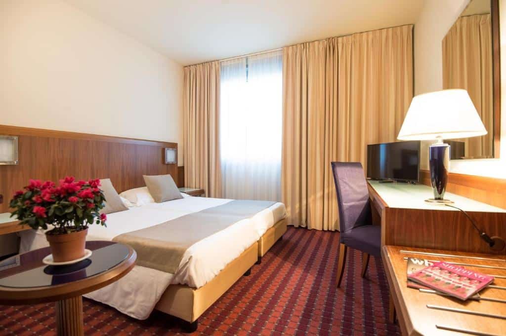 Quarto do First Hotel Malpensa com uma janela com cortinas, uma cama de casal, há uma cabeceira com luminárias presas, há uma mesa de escritório com uma cadeira e a televisão está sob esse móvel, o chão é de carpete vermelho e laranja, para representar hotéis perto do aeroporto de Milão