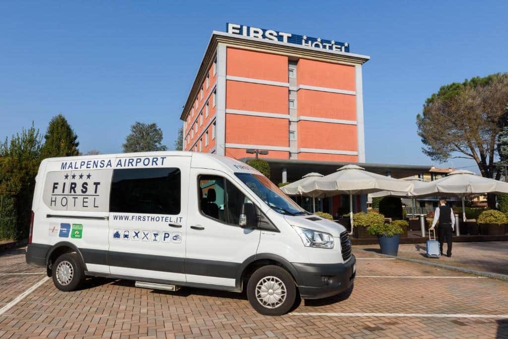 Entrada do First Hotel Malpensa, o prédio é laranja com branco, há vagas na frente da propriedade, além de vasos de plantas e guarda-sóis, em destaque na imagem, há uma van responsável pelo transfer dos viajantes oferecida pelo hotel