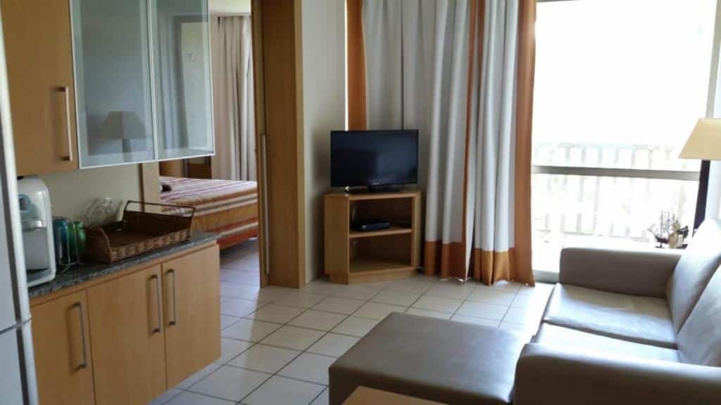 Uma área de estar compacta apresenta um sofá, TV em um suporte e uma cozinha compacta. Uma cama é visível em um quarto adjacente com cortinas no Flat Beach Class Muro Alto em Porto de Galinhas