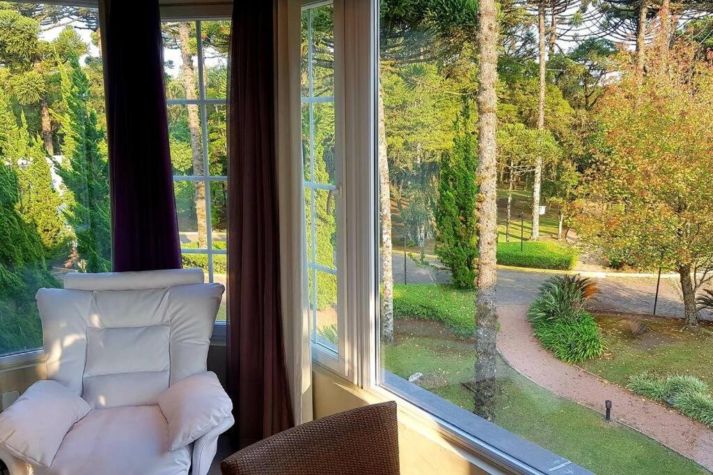 A imagem retrata um flat no Flat no Laje de Pedra no Mountain Village. Há uma janela extensa com vista para a arborização. Uma poltrona está à esquerda, e há uma cadeira com mesa ao seu lado, em uma parte não visível na foto.