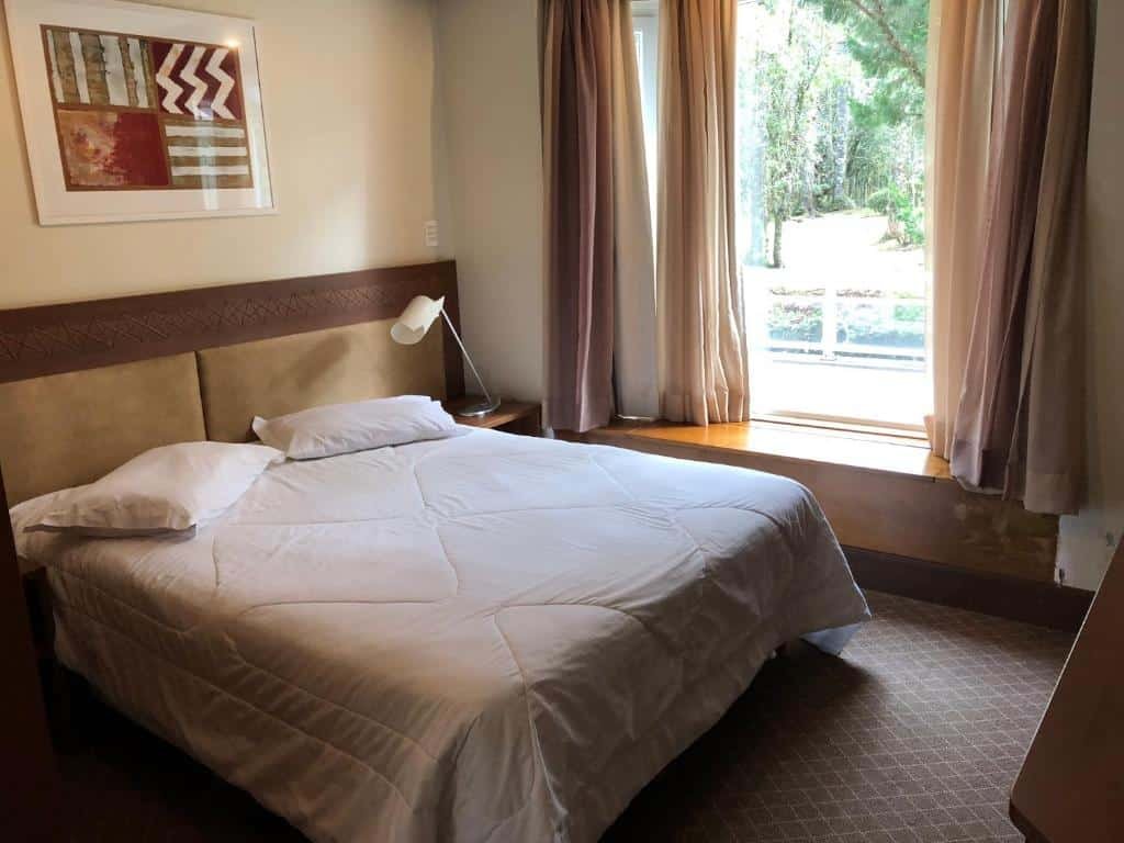 Foto do quarto Flat Luxo Mountain Village. Há uma cama box de casal uma cabeceira atrás dela e uma mesa de cabeceira com abajur no seu lado direito. Uma janela está à direita da cama. Representa o post sobre airbnb em Canela.