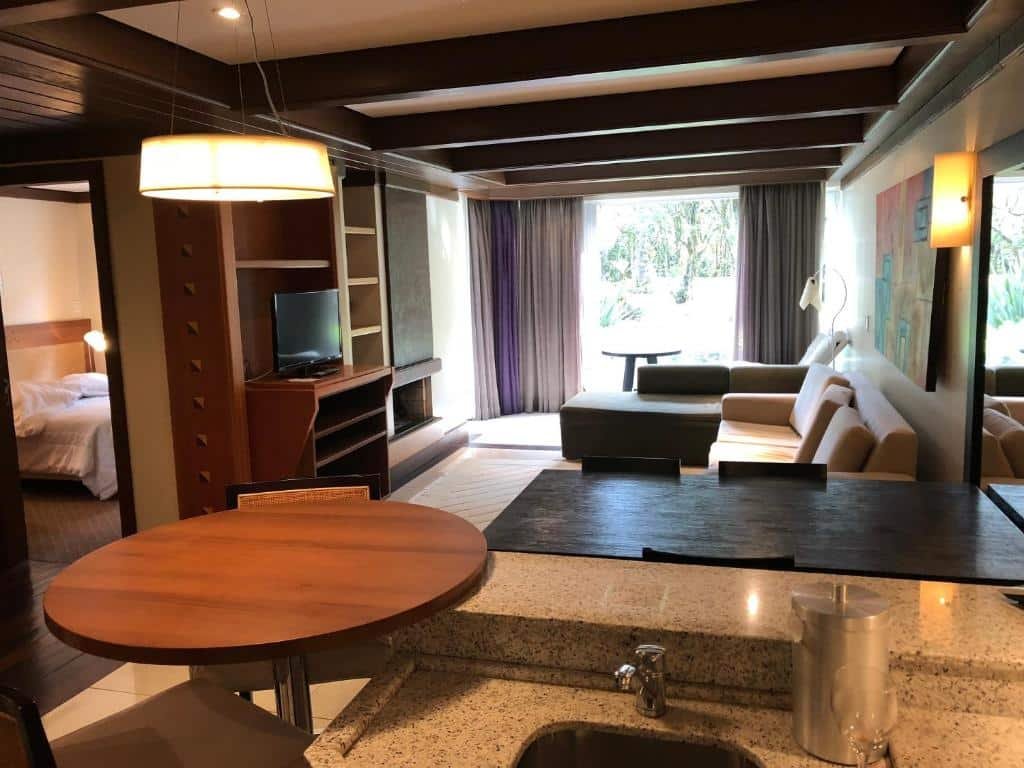 Foto mostrando a sala e parte da cozinha do Flat Luxo Mountain Village. No canto inferior direito vemos parte da pia, e do esquerdo, de uma mesa. No fundo está a sala com sofá à direita e um rack de televisão à esquerda, do lado da porta do quarto.