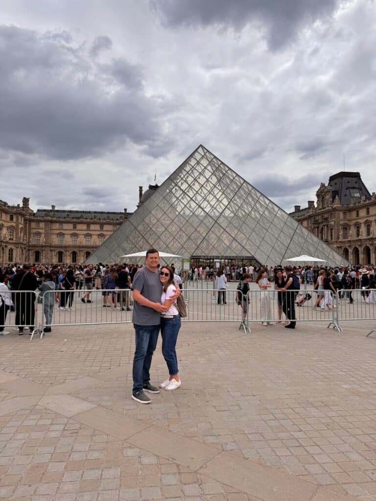 Um casal está em frente à entrada da pirâmide de vidro do Museu do Louvre, em Paris, cercado por outros visitantes.