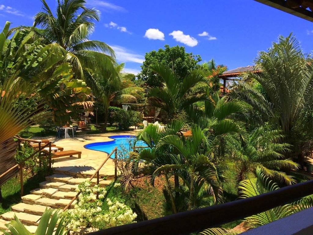 Área da piscina com área verde ao entorno da Pousada Flores do Cerrado, dia de céu azul com algumas nuvens. 