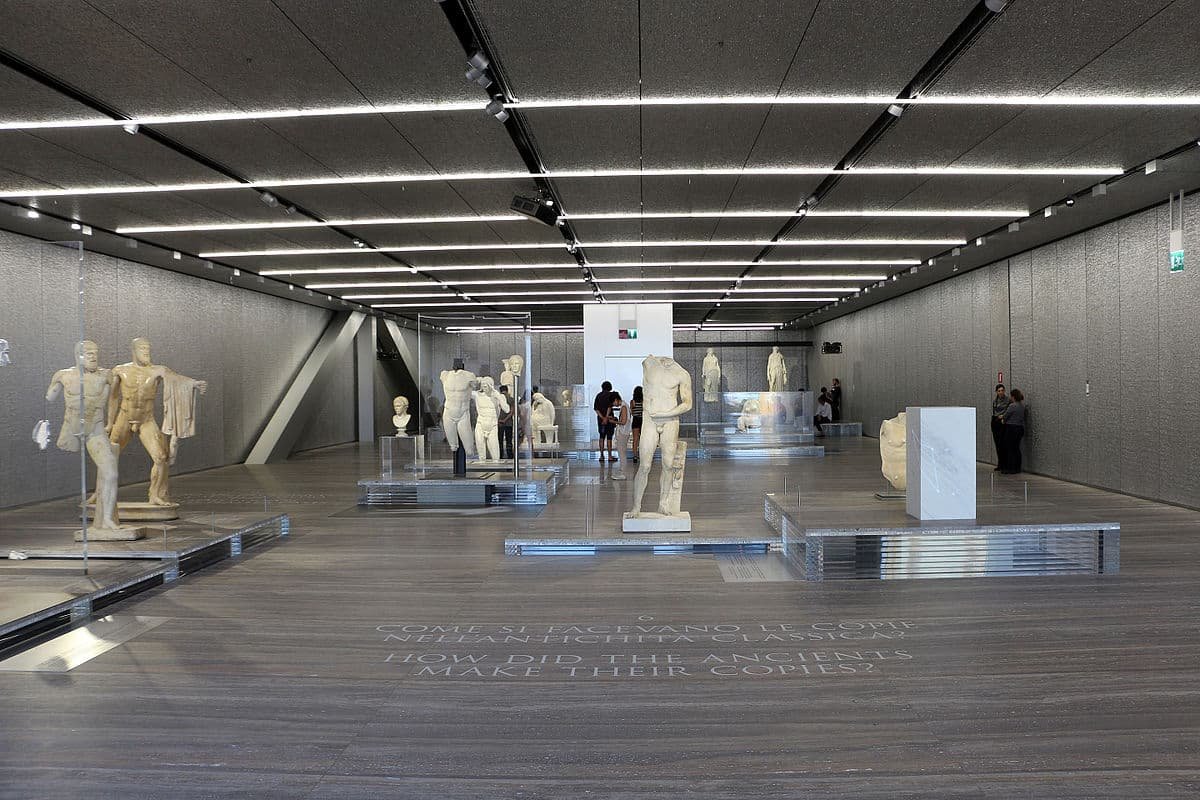 Uma das salas na Fondazione Prada. A galeria exibe diversas esculturas antigas, incluindo bustos e figuras inteiras, sob luzes brilhantes no teto e piso de madeira. É uma boa alternativa de o que fazer em Milão.