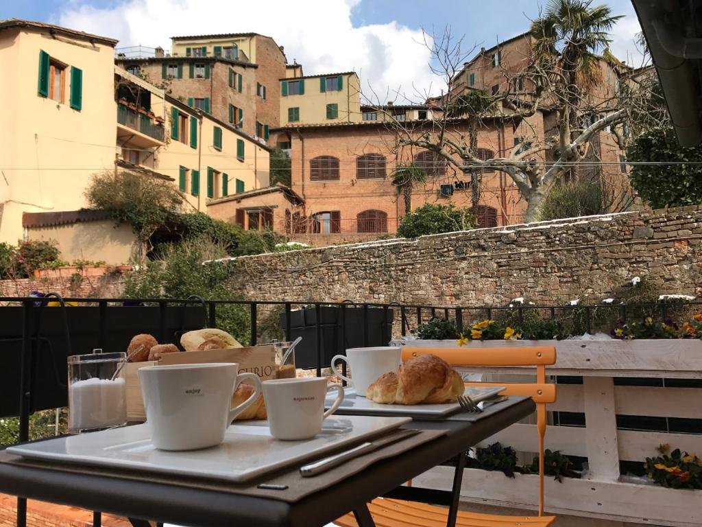 Mesa com xícaras e pães na varanda de um dos hotéis na Toscana, com vista para outros prédios alaranjados da região e muro de tijolos