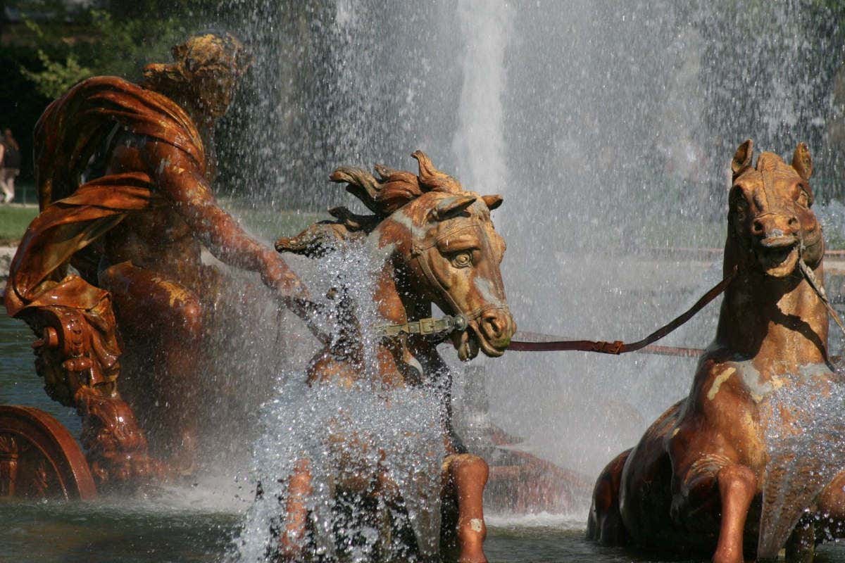 Escultura de fonte no Palácio de Versalhes representando uma figura mitológica com cavalos, cercada por respingos de água.