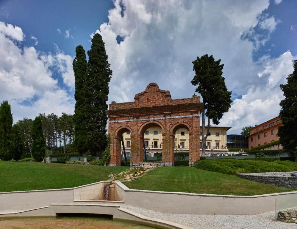 Entrada do Fonteverde Lifestyle & Thermal Retreat, um dos hotéis na Toscana, com portal alto de tijolinhos, bastante vegetação, sendo um gramado verde baixo e árvores altas, e prédios do hotel atrás, sendo um bege e outro laranja