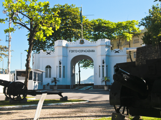 Uma entrada em arco branco com a inscrição "Forte de Copacabana" com canhões militares em primeiro plano e árvores ao redor da estrutura.