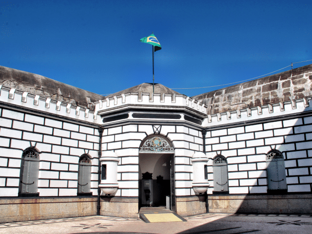 Fortaleza branca com uma bandeira brasileira no topo, portas e janelas em arco sob um céu azul claro.