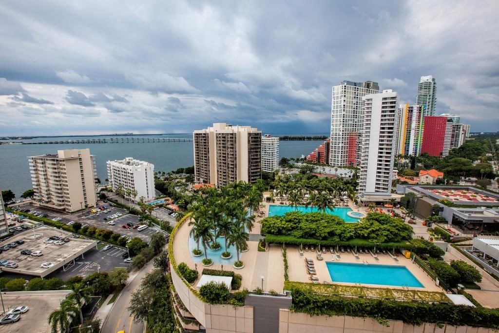 A imagem mostra uma vista aérea de uma área urbana com vários edifícios altos, alguns coloridos, e uma grande área verde. No centro, há duas grandes piscinas rodeadas por palmeiras e espreguiçadeiras. À direita, há mais edifícios altos e modernos. Ao fundo, pode-se ver a baía com uma longa ponte que a atravessa, sob um céu parcialmente nublado. Há também áreas de estacionamento e ruas arborizadas ao redor dos edifícios. A cena combina elementos naturais e urbanos, com um foco em áreas de lazer e relaxamento do Fortune House Hotel, um dos hotéis em Miami Beach indicados.